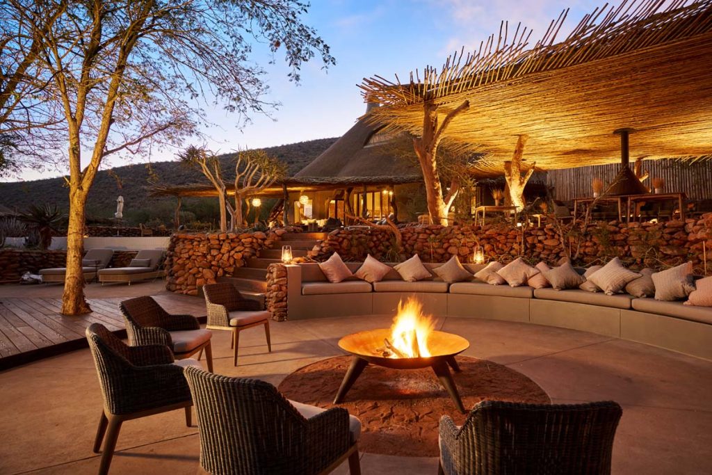 A small fire pit with armchairs on a stone terrace. Straw bale roof building is in the back.