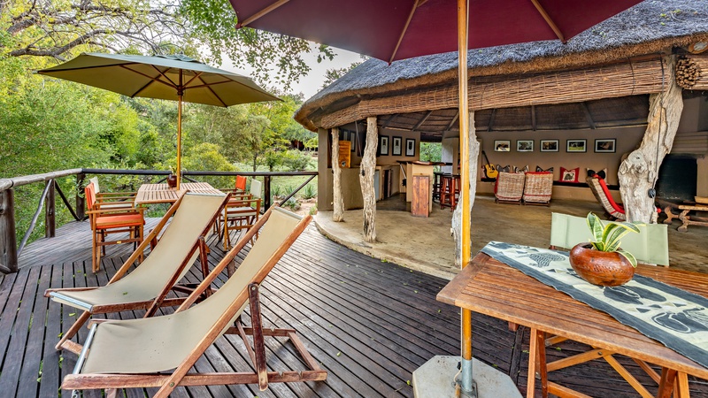 A wooden balcony with chairs and umbrella. There is a wooden round hut at the back completely open, no door. 