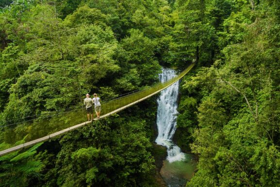A couple is standing on a hanging bridge overlooking a waterfall