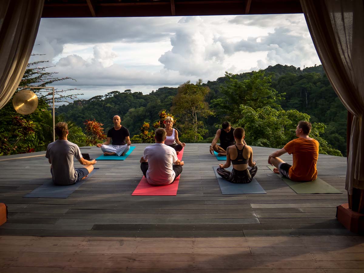 Several people doing yoga on a wooden platform overlooking rainforest and ocean