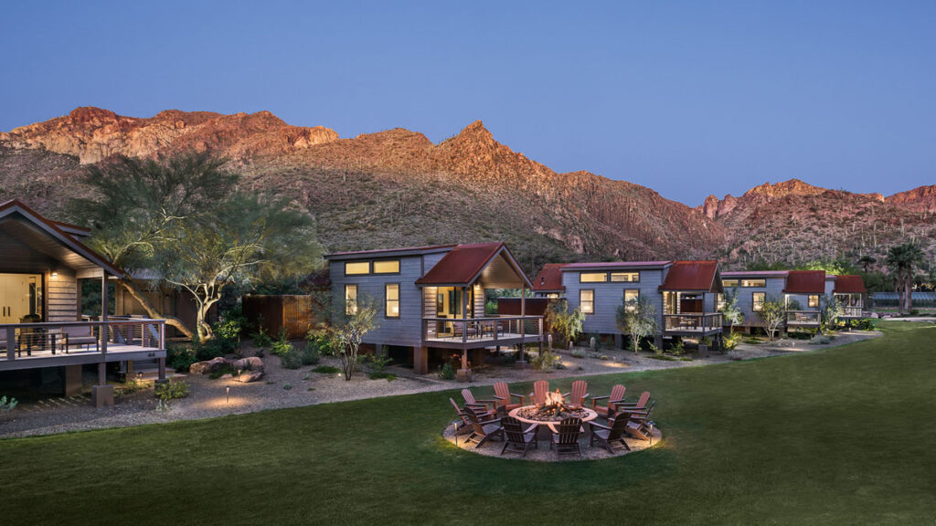 Several wooden cottages in front of a mountain. Green garden and a fire pit with chairs around are in front.