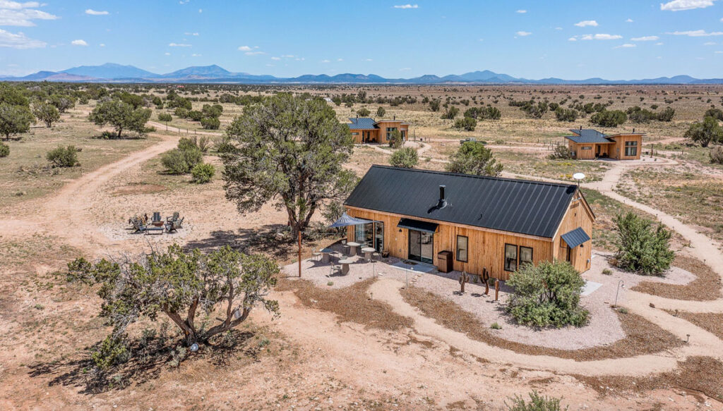 A wooden house with black roof in a middle of a dry sandy land with only a handful of trees and bushes.