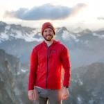 A man with grey jeans, red jumper and red hat is standing and is surrounded by mountains.