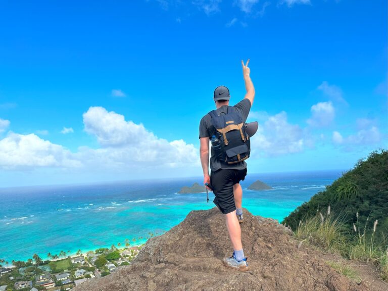 A man is standing on a cliff with one hand up high overlooking turquoise-colored ocean.