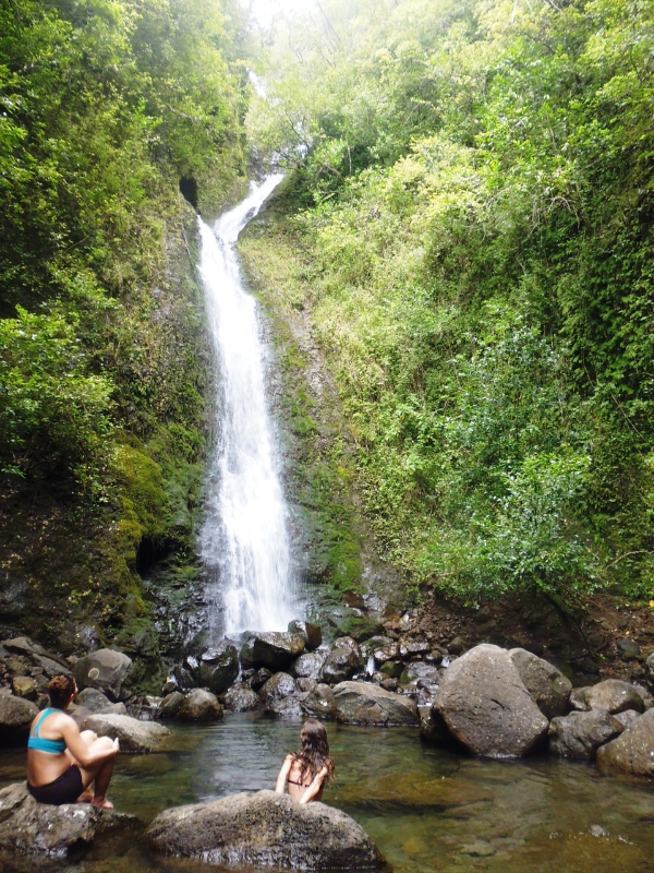 A medium range waterfall in lush jungle. Two people are sitting on the rocks below and watching it.