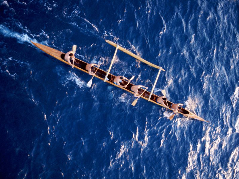 An outrigger canoe with 6 people on the blue ocean from above