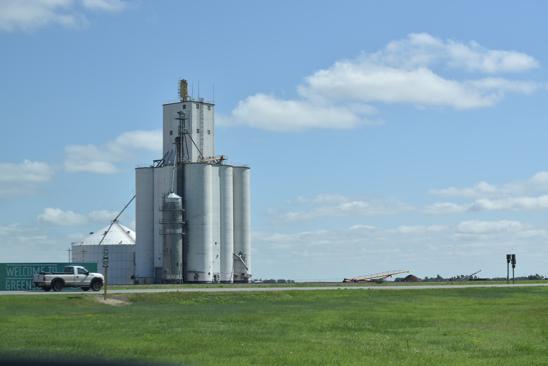 A large grain silo and a pick-up truck on a large green field. 