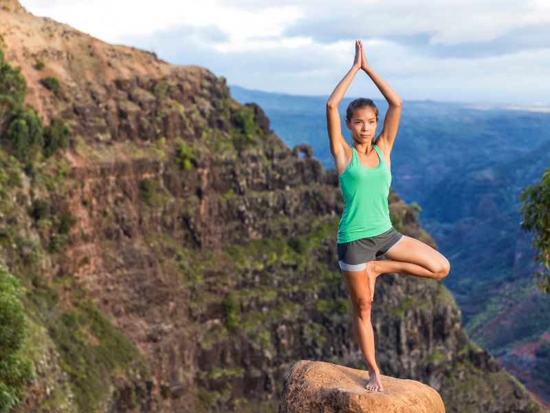 A woman in grey shorts and a green shirt stands on one leg striking a yoga pose on top of a cliff with a rocky mountain behind.