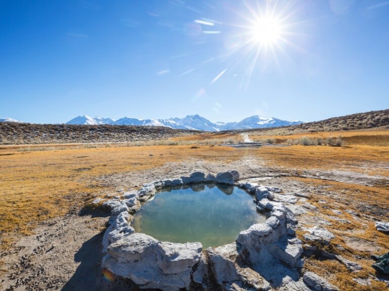 A small natural pool surrounded by grey rocks with an open pasture and snowy mountains as backdrop.