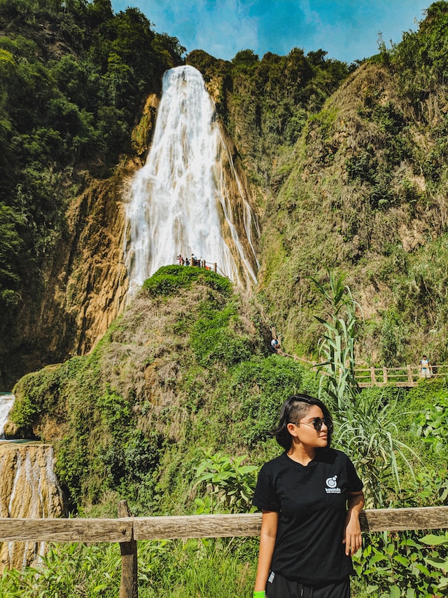 A woman in black T-shirt is standing in front of a waterfall and a green hill with more people on it. 