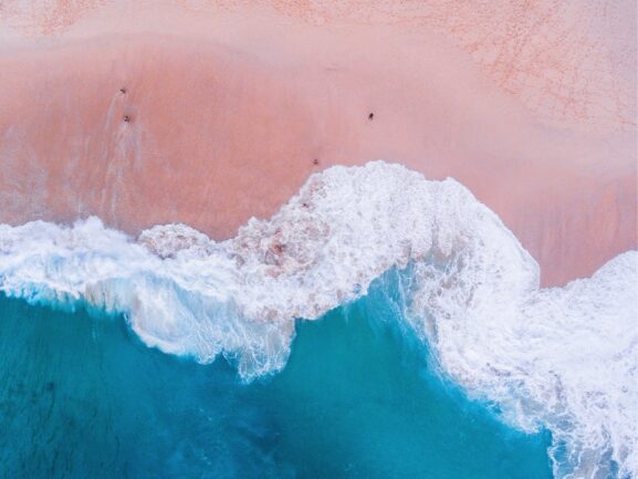 A few swimmers on a pink beach with white waves and turquoise water from above.