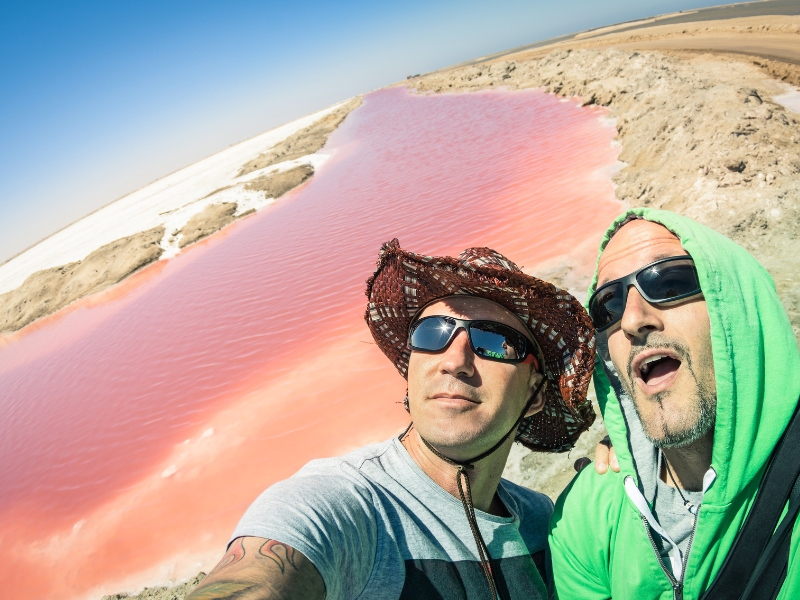 Two men is taking a selfie in front of a pink lake. 