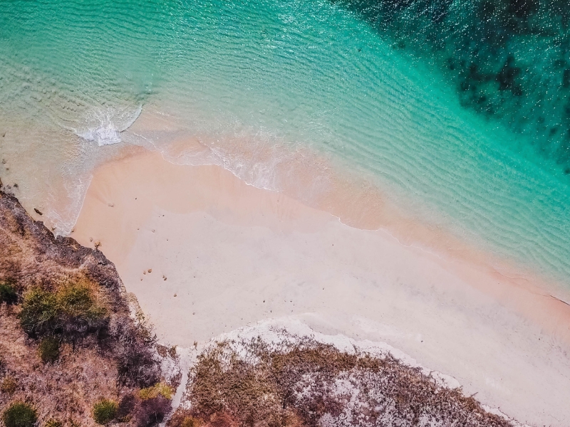 Pink sandy beach from above with green ocean and rocks.