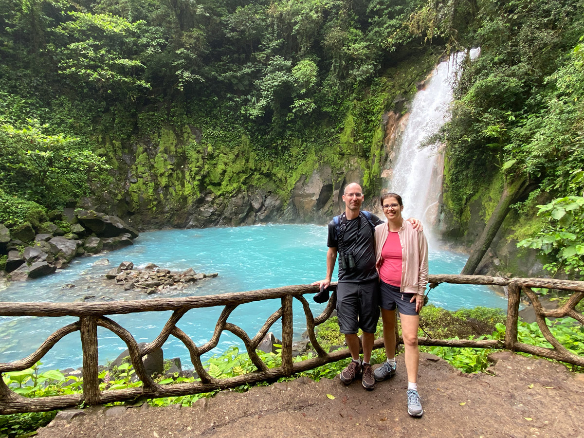 A man and a woman in front of a turquoise lake and waterfall.