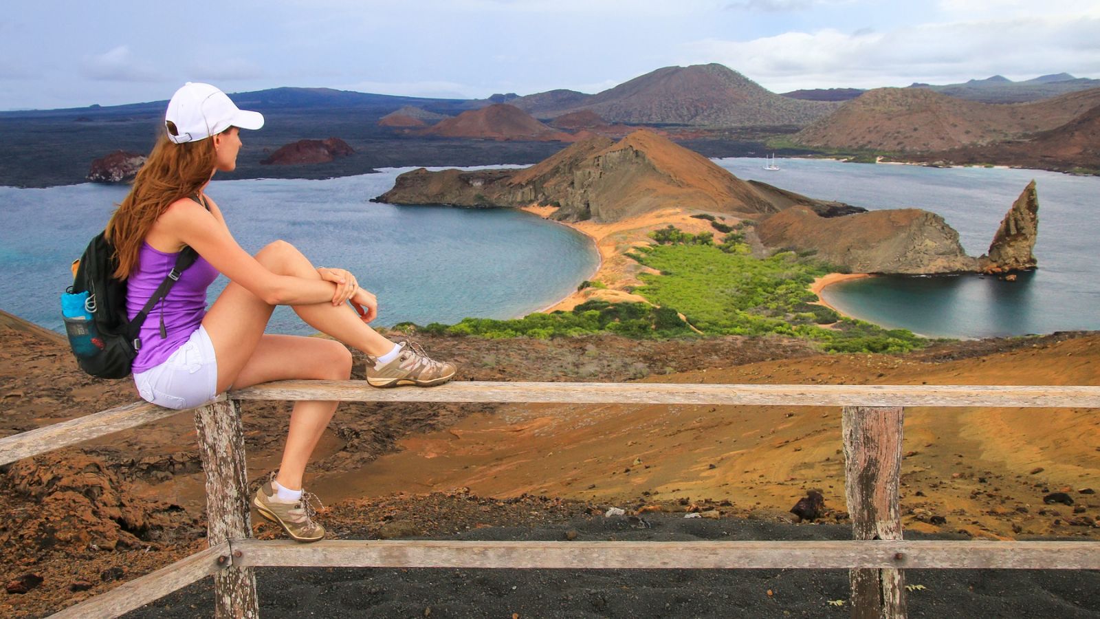 A woman with purple shirt and white shorts looking at a cliff formation and two beaches in the Galapagos islands.
